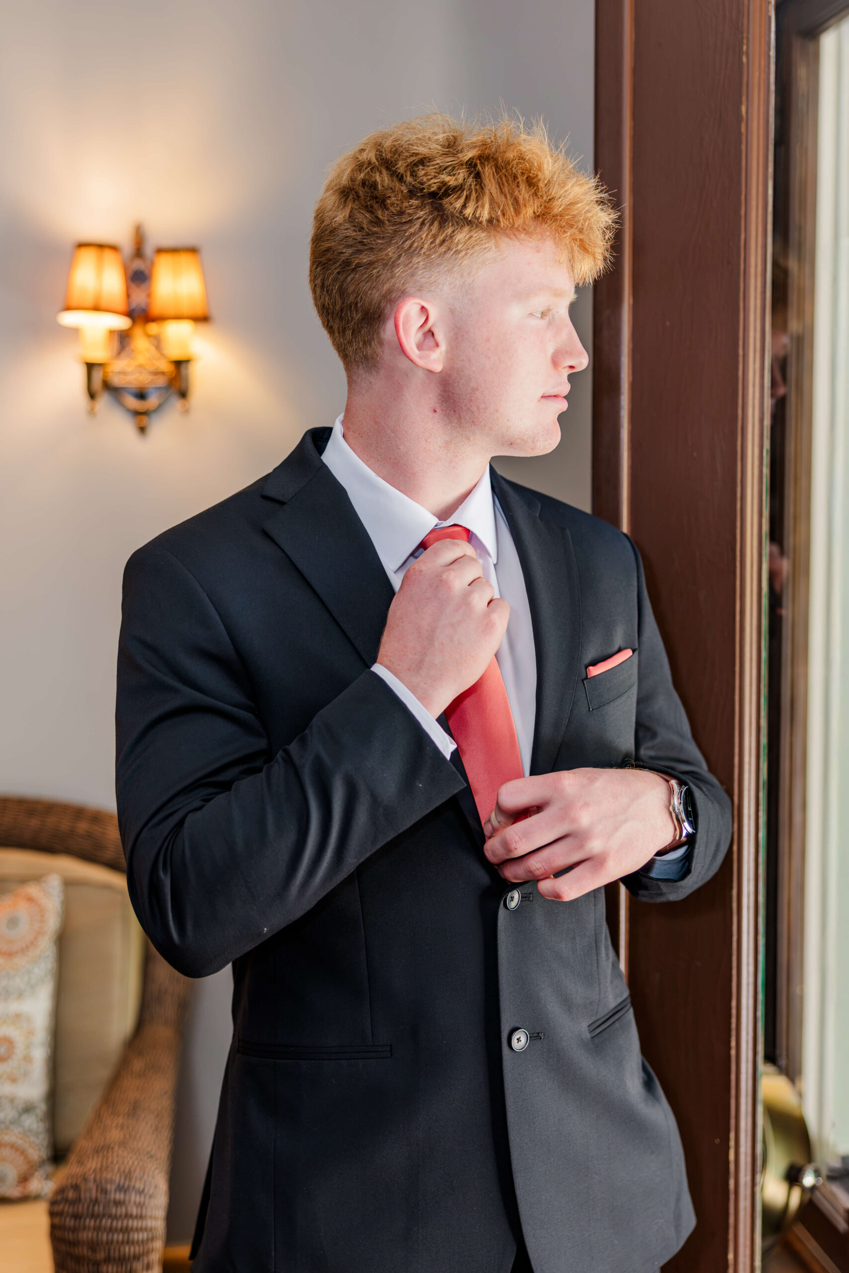 A groom adjusts his orange tie while gazing out a window before his Royal Park Hotel wedding