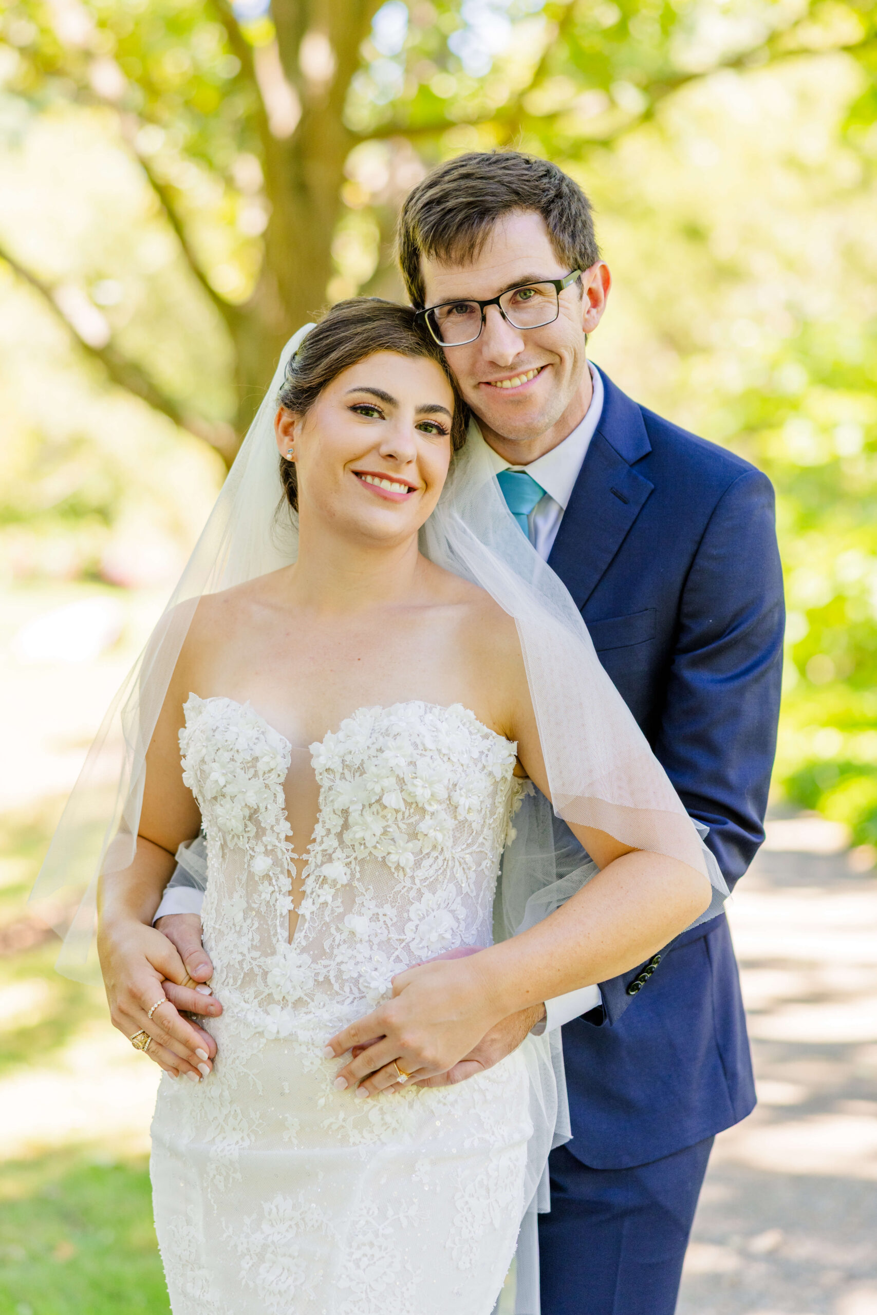 A bride leans into her groom in a blue suit while hugging from behind in a sidewalk