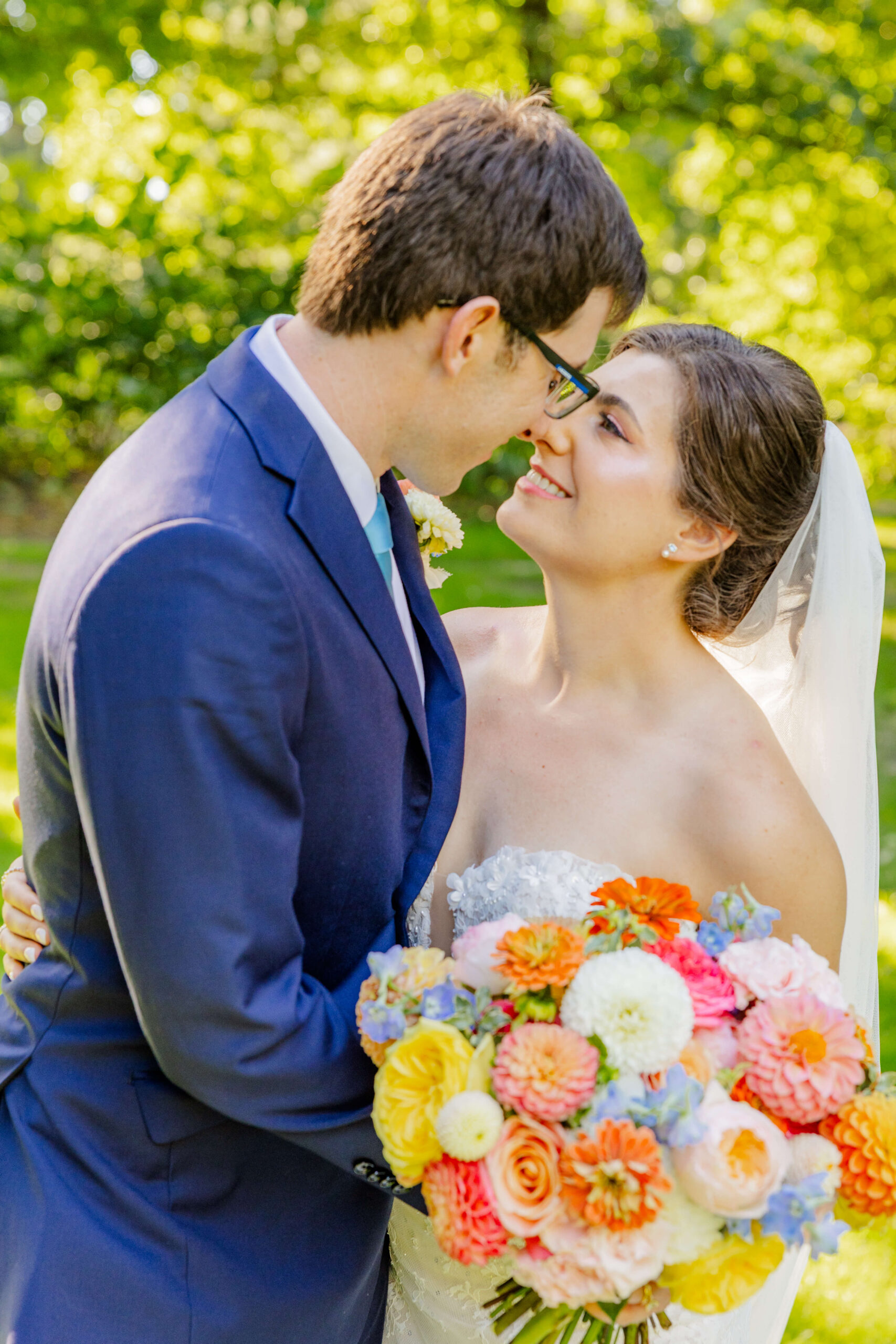A happy bride and her groom in blue lean in for a kiss while standing in a lawn holding a colorful bouquet at their woodbridge event barn wedding